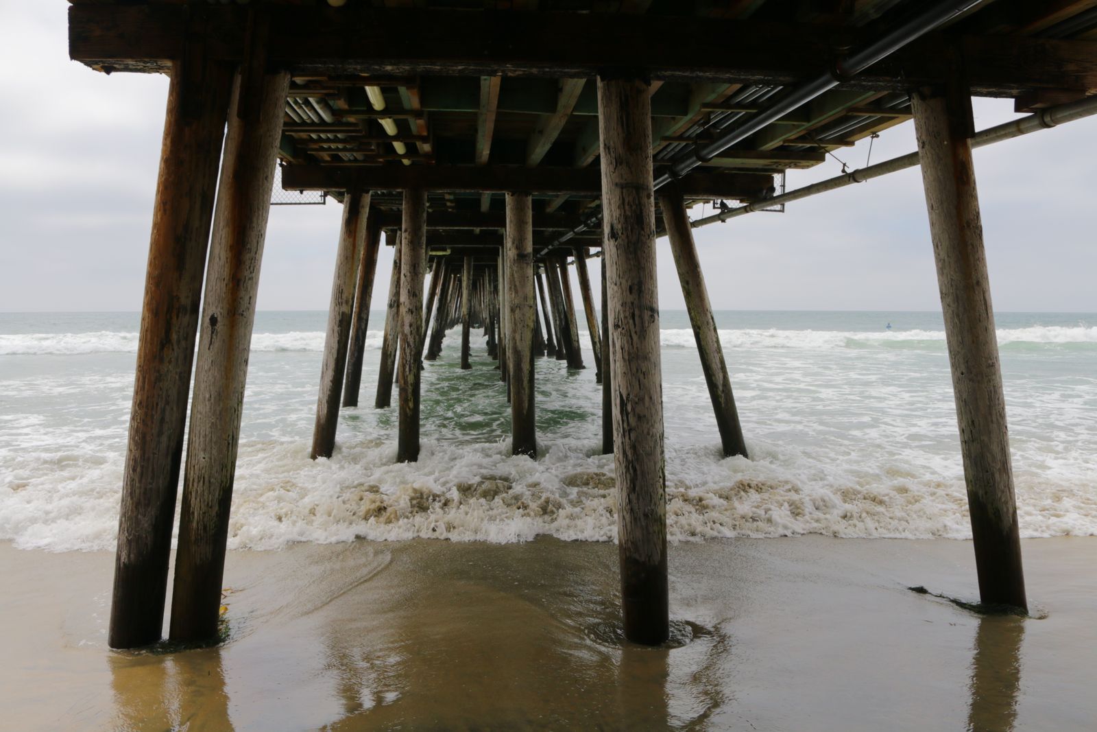 View from beneath a weathered wooden pier with waves crashing on a cloudy seaside