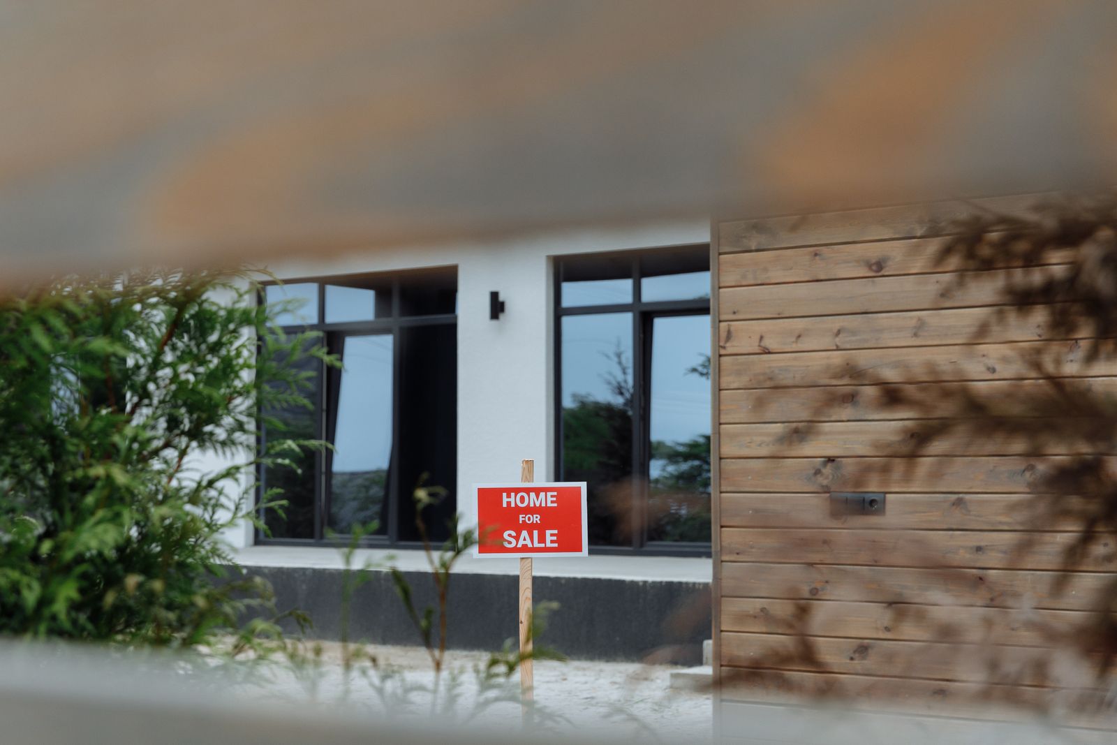 A contemporary home framed by greenery with a For Sale sign in the front yard