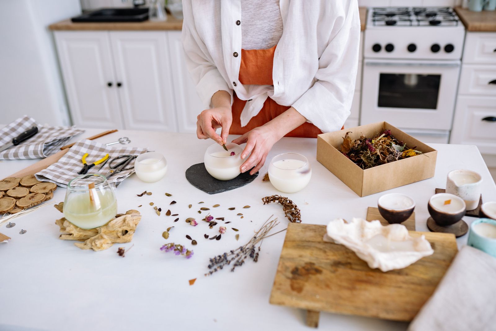 Artisan hand-pouring a candle amid dried flowers in a cozy candle-making workshop